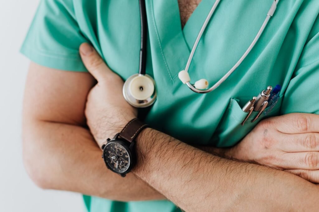 Unrecognizable man wearing medical uniform with stethoscope and pens standing in hospital with crossed arms during work in day time